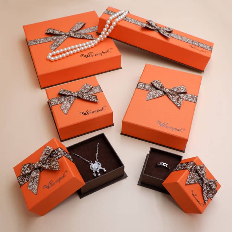Collection of luxury custom orange jewelry packaging boxes with brown ribbon bows, showing an open pendant necklace box, a ring box, and draped pearls.