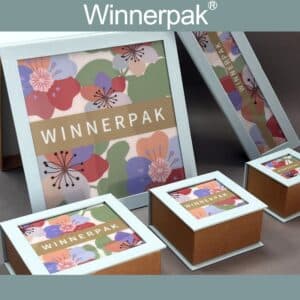 Top-angle view of a wholesale cardboard jewelry box from the BGLP collection, featuring a textured light blue border, brown base, and a floral printed insert visible through a clear PVC top window.